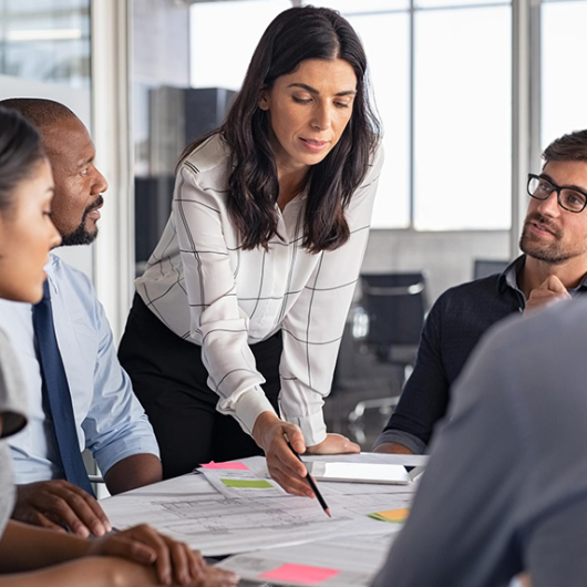 Group of professionals in a meeting room, with a woman leading the discussion by pointing at documents on the table.